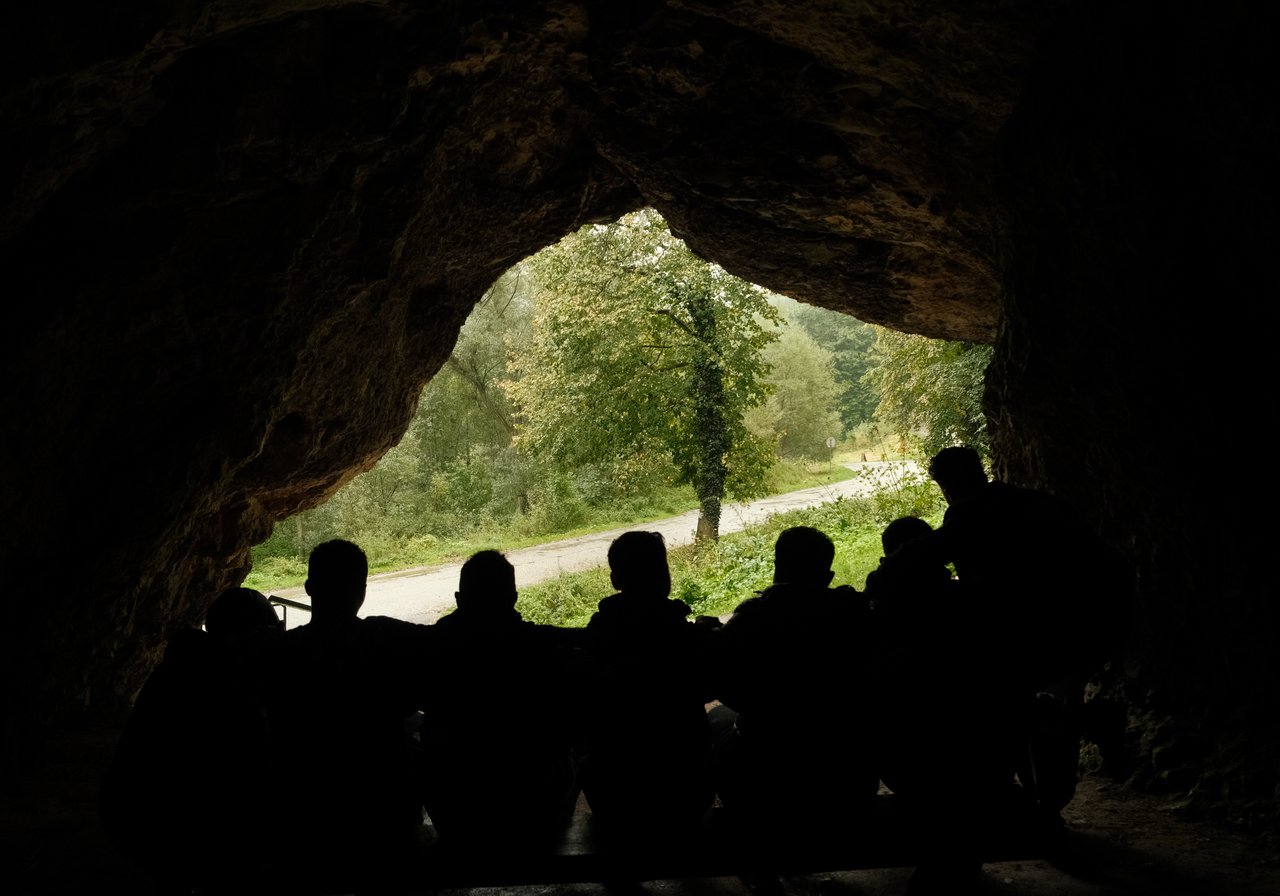 Picture shot from inside of a cave showing the silhouette of men standing arm in arm looking out of the cave
